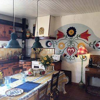 Eetkeuken met geel-wit geblokt tafelkleed, blauwe placemats, felgele bloemen en kleurrijke muurschildering in Zuid-Franse stijl